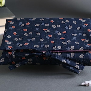 May include: A navy blue quilted fabric case with a repeating pattern of white, orange, and gray leaf shapes. The case is tied with a matching fabric bow. The case is likely for storing items.