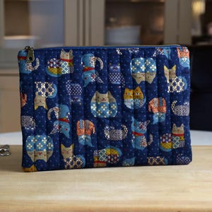 Peut inclure: Une pochette matelassée rectangulaire en tissu bleu marine profond ornée d'un motif répétitif de chats stylisés aux imprimés géométriques et floraux variés. La pochette comprend une fermeture éclair en métal sur le bord supérieur.
