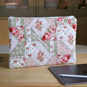 May include: A rectangular quilted pouch featuring a patchwork design of floral patterns in shades of pink, green, and white. The fabric displays various motifs including rose bouquets, small floral sprigs, and green stripes, finished with a white zipper.