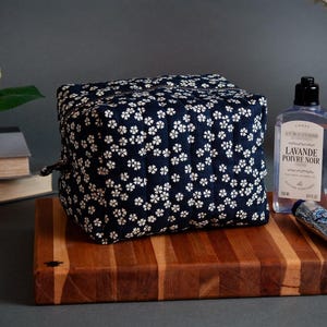 Könnte beinhalten: Eine marineblaue Kosmetiktasche mit weißem Blumenmuster liegt auf einem Holzschneidebrett. Daneben befinden sich Schönheitsprodukte, darunter eine Flasche Lavendel-Lotion und eine kleine Tube Handcreme.