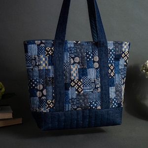 Puede incluir: Un bolso tote grande con un diseño de patchwork en tonos azules y crema. El bolso tiene asas azules resistentes y una base acolchada. El fondo incluye libros, un jarrón con flores blancas y una planta verde.