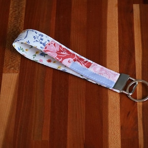 May include: A fabric wristlet keychain featuring a white background with a floral pattern in shades of red, pink, blue, and yellow. The strap is secured with a silver-toned metal clamp and includes a circular metal split ring for keys.
