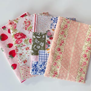 May include: Five fabric items with various patterns, including strawberries, roses, and patchwork designs. The items are in shades of pink, red, white, and green. The items appear to be quilted or padded.