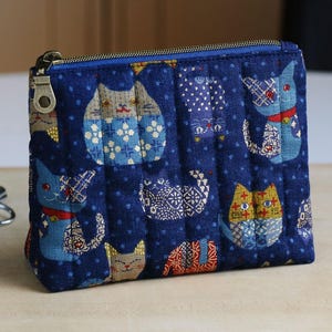 May include: A rectangular quilted fabric pouch in a deep navy blue color featuring a repeating pattern of stylized cats in various colors and geometric prints. The pouch includes a blue zipper closure with a metallic pull tab.