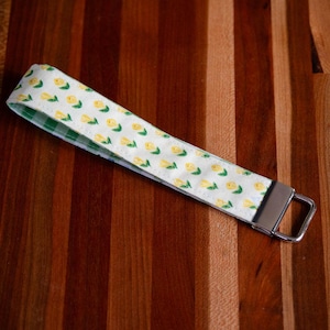 May include: A white wristlet key fob with a yellow tulip pattern and a green gingham lining. The key fob has a silver-colored metal clasp and is laying on a wooden surface.