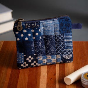 May include: A small, square, patchwork coin purse in shades of blue with a brass zipper. The purse features various geometric patterns and a small blue fabric loop. A white cylindrical object and a small metal tin sit on a wooden surface.