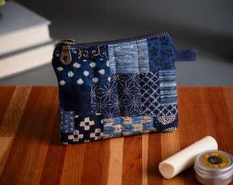 Op de afbeelding: Een kleine, vierkante patchwork-muntportemonnee in blauwtinten met een messing rits. De portemonnee heeft verschillende geometrische patronen en een kleine blauwe stoffen lus. Een wit cilindrisch object en een kleine metalen doos liggen op een houten oppervlak.