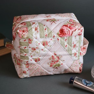 Könnte beinhalten: Ein quadratischer, gesteppter Kosmetikbeutel mit floralem Muster. Die Tasche hat ein Patchwork-Design mit rosa Rosen, grünen Streifen und kleinen Blumenprints. Sie hat einen Reißverschluss und kleine Stoffgriffe.