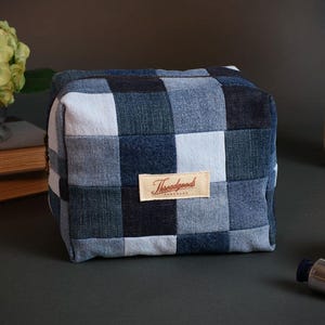 May include: A square, denim-style cosmetic bag with a patchwork design in shades of blue and white. The bag features a rectangular label with the text "Threadgoods". The bag is sitting on a dark surface.