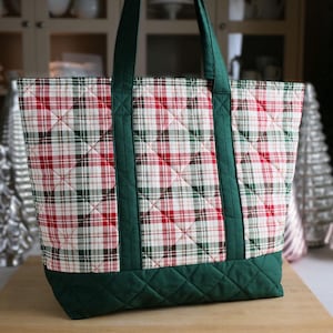 May include: A quilted tote bag featuring a red, green, and white plaid pattern on the main body. The bag includes solid dark green fabric panels at the base and along the vertical straps, which extend upward to form two handles for carrying.