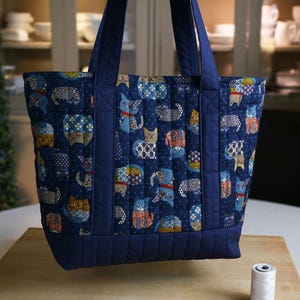 May include: A quilted tote bag featuring a dark blue background with a repeating pattern of stylized cats in various colors and geometric designs. The bag has solid dark blue fabric panels at the base and top, with matching dark blue shoulder straps.