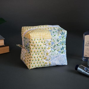May include: A patchwork cosmetic bag with a yellow gingham and floral pattern. The bag has a zipper closure and a small loop handle. Other items in the image include a bottle of lotion, a jar, a tube of cream, and books.
