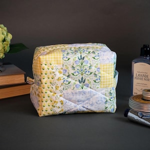 May include: A quilted cosmetic bag with a patchwork design in yellow and blue floral patterns. The bag is square-shaped with a zipper closure. Other items include a bottle of lotion, a jar, and a tube of cream.