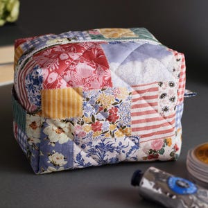 May include: A cube-shaped, quilted fabric pouch with a patchwork design. The patchwork features various floral, striped, and patterned fabrics in red, blue, yellow, and white. The pouch has a zipper closure and a small fabric loop.