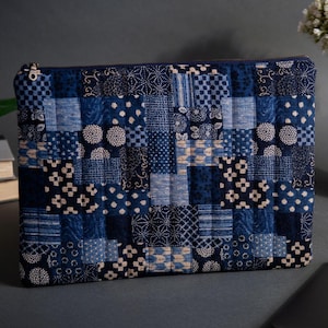 Peut inclure: Une pochette rectangulaire matelassée avec un motif patchwork dans les tons de bleu et de blanc cassé. Le motif comprend divers motifs géométriques et floraux. Une fermeture éclair dorée longe le bord supérieur. La pochette est probablement utilisée pour transporter de petits objets.