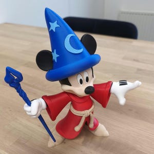 Könnte beinhalten: Eine Mickey Mouse-Figur als Zauberer, mit blauem Hut mit Sternen und Mond, rotem Umhang, weißen Handschuhen und blauem Stab. Die Figur steht auf einer Holzoberfläche.