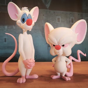May include: Two white mouse figurines with pink ears, tails, and feet. One mouse has a red nose and blue eyes, the other has a stern expression. The figurines are standing on a wooden surface.