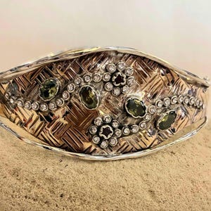 Könnte beinhalten: Ein silber- und goldfarbenes Armband mit gewebter Textur. Das Armband ist mit grünen Edelsteinen und kleinen, klaren Kristallen in floralen Mustern verziert. Das Design ist aufwendig und detailliert, passend für besondere Anlässe.