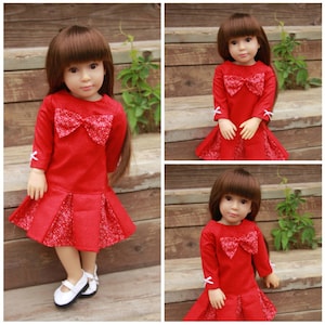 Puede incluir: Un vestido rojo con un detalle de lazo blanco y mangas largas. El vestido tiene una falda plisada y lo lleva una muñeca con el pelo castaño.