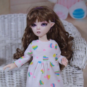 May include: A doll with long brown curly hair wearing a white dress with rainbow hearts. The doll has purple eyes and pointy ears.
