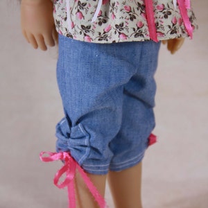 May include: A doll wearing a floral print top with pink ribbons and blue denim shorts with pink ribbons tied at the bottom.