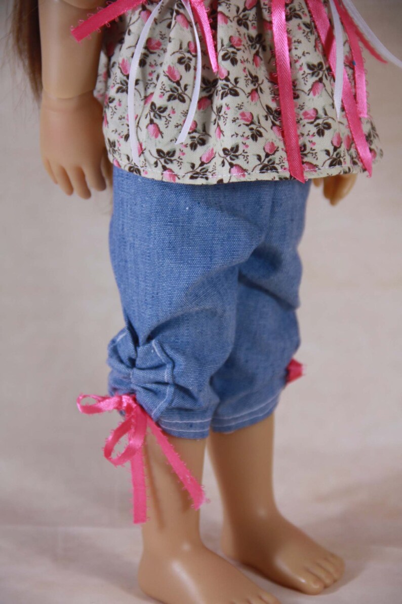 May include: A doll wearing a floral print top with pink ribbons and blue denim shorts with pink ribbons tied at the bottom.