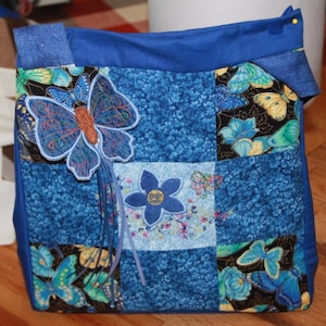 May include: A blue patchwork tote bag featuring a butterfly design. The bag has a blue fabric strap and is decorated with embroidered butterflies and flowers.