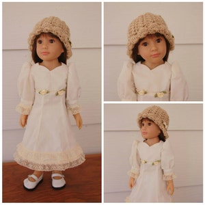 以下が含まれることがあります： レースのトリムが付いた白いドレスと、かぎ針編みの帽子をかぶった人形。人形は茶色の髪と青い目です。