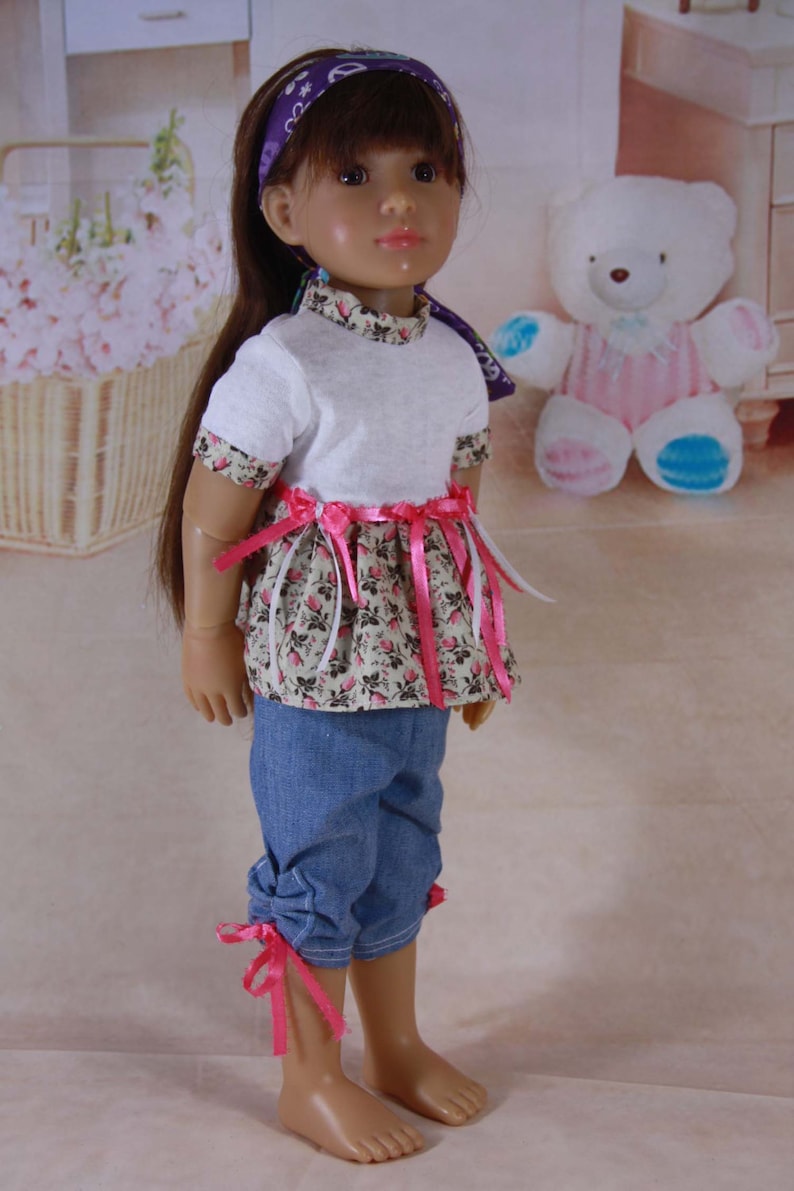 May include: A doll with long brown hair wearing a white t-shirt with a floral print skirt and denim pants with pink ribbon ties. The doll is wearing a purple headband with a peace sign design.