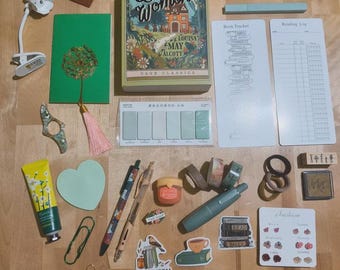 May include: An assortment of stationery items, including a copy of "Little Women," pens, stickers, washi tape, and a reading light. The items are arranged on a wooden surface, with a green journal and heart-shaped accents.
