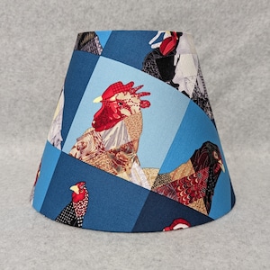 May include: A blue lampshade with a patchwork design featuring roosters and hens. The fabric is a mix of blue, red, white, and brown.