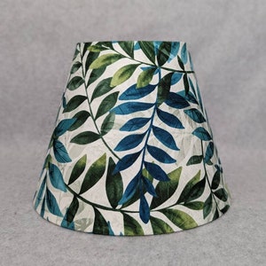 May include: A tapered lampshade featuring a botanical leaf pattern. The leaves are in shades of green and teal against a white background. The lampshade is designed to fit on a lamp base and provide ambient lighting.