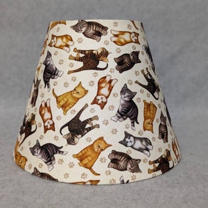 May include: A cream-colored lampshade with a pattern of playful cats in various poses. The cats are brown, orange, and gray with black stripes. The background is a light beige with a paw print pattern.