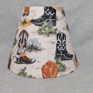 May include: A beige lampshade with a repeating pattern of cowboy boots, hats, and cacti. The boots are in various colors, including black, brown, and white, with decorative stitching. The cacti are green, and the hats are tan.