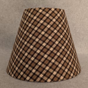 May include: A tapered lampshade with a brown and beige plaid pattern. The fabric shade has a classic design, suitable for a table or floor lamp. The plaid pattern is a repeating grid of intersecting lines.