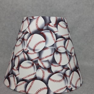 May include: A white lampshade with a baseball pattern. The lampshade is made of fabric and has a conical shape.