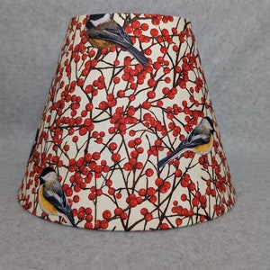 May include: A lampshade with a cream background and a pattern of red berries and brown branches with three small birds. The birds are brown, white, and yellow.