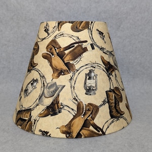 May include: A beige lampshade with a western theme featuring a repeating pattern of cowboy boots, saddles, hats, and barbed wire.