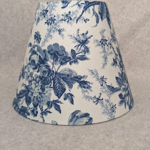 May include: A lampshade with a classic toile pattern in shades of blue and white. The fabric features a detailed floral and botanical design, creating a traditional aesthetic. The lampshade has a tapered shape.