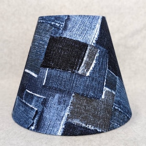 May include: A denim lampshade with a patchwork design. The shade features various shades of blue denim, including lighter and darker washes, arranged in a geometric pattern. The lampshade has a tapered shape.