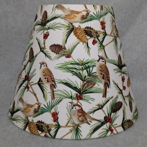 May include: A white lampshade with a repeating pattern of brown sparrows, pine cones, green pine needles, and red berries. The lampshade has a tapered shape, wider at the base and narrower at the top. The design gives a natural, woodland theme.