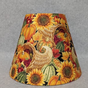 May include: A lampshade with a black background and a colorful autumn harvest design. The design features pumpkins, sunflowers, corn, and fall leaves.