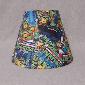 May include: A lampshade featuring a Teenage Mutant Ninja Turtles comic book design. The lampshade is blue, green, and black with images of the turtles and the words "Teenage Mutant Ninja Turtles" and "Adventures".