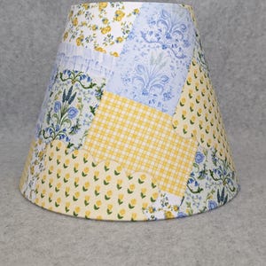 May include: A patchwork lampshade with a variety of floral and patterned fabrics. The design includes yellow gingham, blue floral patterns, and white fabric with yellow flowers. The lampshade has a conical shape.