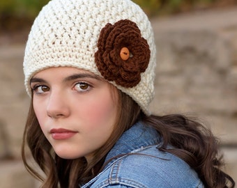May include: A white crocheted beanie with a brown crocheted flower and a brown button in the center. The beanie is worn by a person with long brown hair and a blue denim jacket.