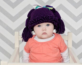 May include: A baby wearing a purple crocheted hat with pigtails. The hat has a bow on top and the baby is sitting in a white chair against a gray and white chevron patterned background.