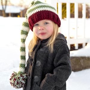 May include: A young person wearing a red, white, and green striped elf hat with a long, pointed tip. The hat has a pom-pom at the end of the tip. The person is wearing a dark gray coat and is standing in the snow.