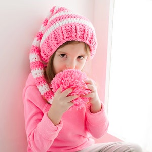 May include: A young girl wearing a pink and white striped crocheted hat and matching scarf holds a pink pom-pom in her hands.