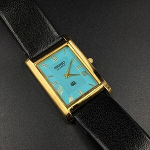 Könnte beinhalten: Eine rechteckige Seiko Quartz Armbanduhr mit goldfarbenem Gehäuse, einem leuchtend türkisfarbenen Zifferblatt mit dezenter Wellenstruktur und goldenen römischen Ziffern. Die Uhr hat goldene Zeiger und ein schwarzes, strukturiertes Lederarmband.