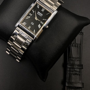 Puede incluir: Un reloj Seiko Quartz rectangular con esfera negra y correa de metal plateado. El reloj presenta indicadores de hora con números romanos y un pequeño subdial de segundos. También hay una correa de reloj de cuero negro adicional.